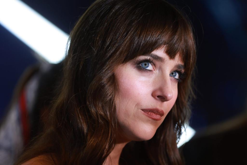 Dakota Johnson looks on during the red carpet for the movie "Madame Web" close up shot wearing red lips and dark brown hair with fringe.